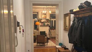 Ein Zimmer mit Holzdielen im Fischgrätmuster, einem Holztisch, einem Stuhl, einem Kleiderständer mit Jacken und einem Stativ mit einer Kamera.