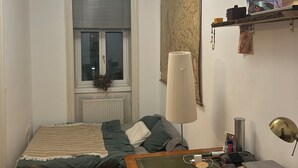 Ein Schlafzimmer mit einem Bett, einem Schreibtisch mit einem Laptop, einem Stuhl, einer Lampe und einem Fenster mit Jalousien.