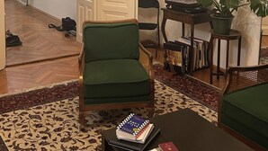 Ein Wohnzimmer mit einem grünen Sessel, einem Couchtisch mit Büchern, einem Bücherregal, einer Topfpflanze und einer Tür zu einem anderen Raum.