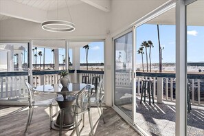 Ein Balkon mit Glastisch und Stühlen, der auf einen Strand mit Palmen und einem Steg blickt.