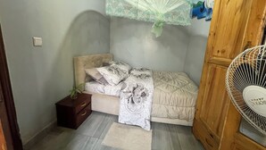 Une petite chambre à coucher, bien rangée, avec un lit, une table de chevet et un ventilateur sur pied.