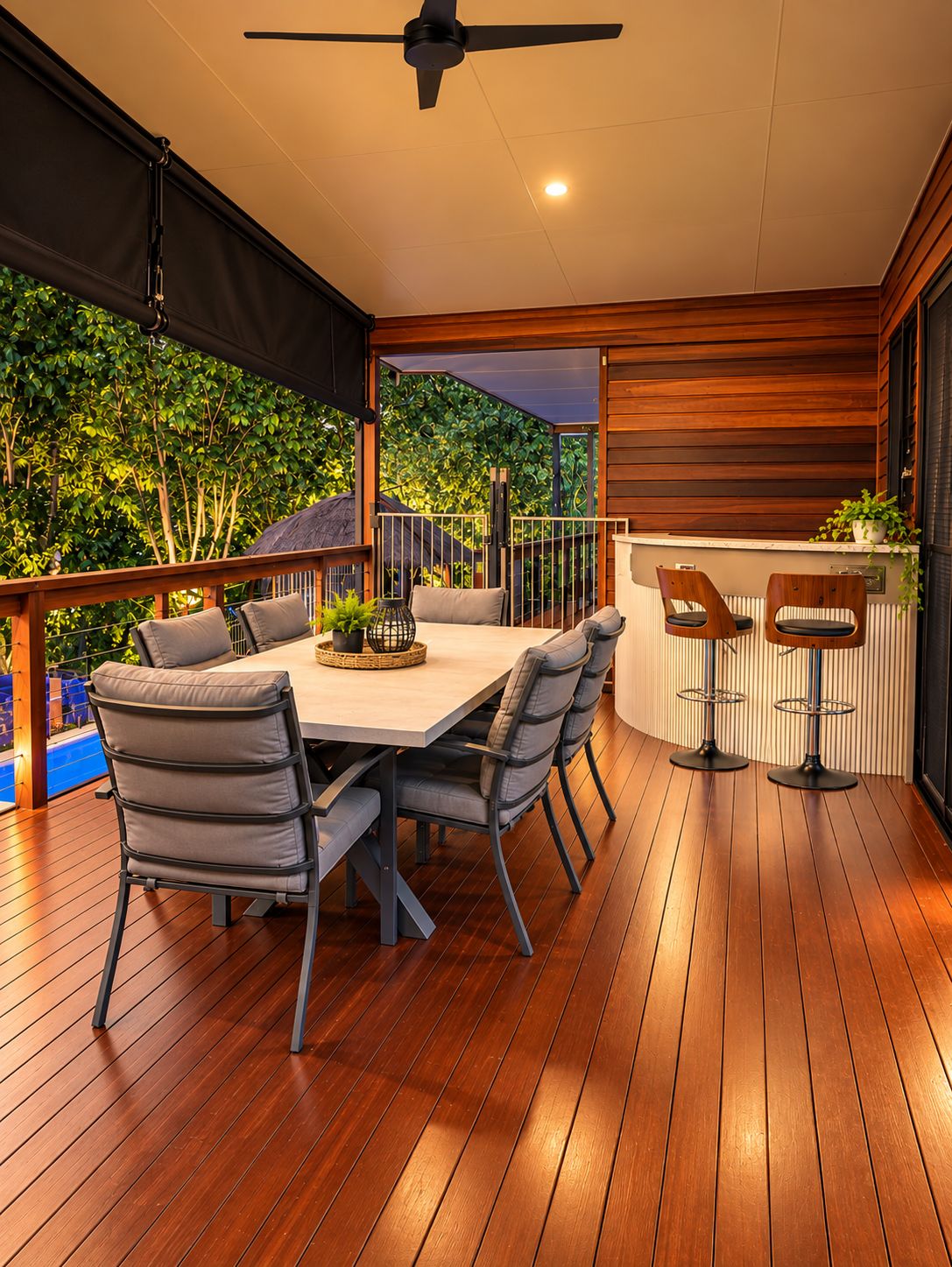 An outdoor patio with a wooden table and chairs, a ceiling fan, and a view of greenery.