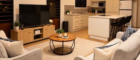 A modern living room with a flat-screen TV, a ceiling fan, and a kitchen area with a microwave and refrigerator.