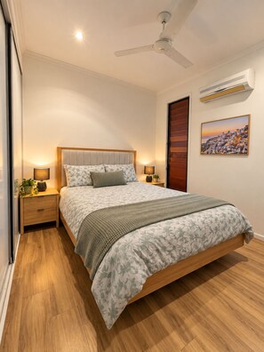 A bedroom with a bed, nightstands, a ceiling fan, an air conditioning unit, and a painting on the wall.