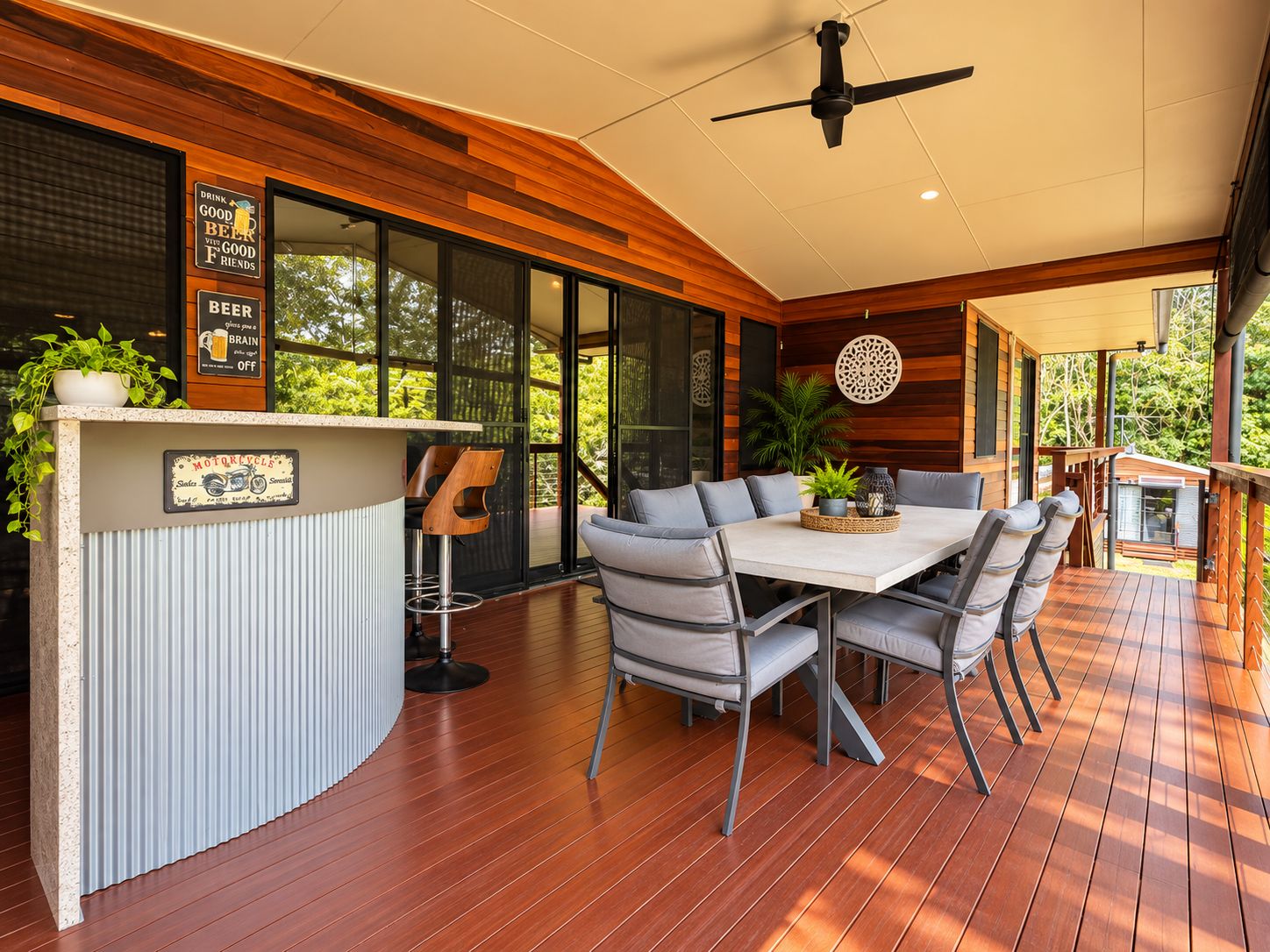 A modern outdoor dining area with a wooden deck, a bar counter, and a ceiling fan.