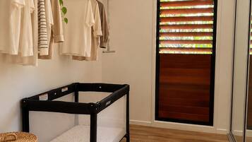 A room with a crib, a clothing rack, a basket, a woven basket, and a window with blinds.