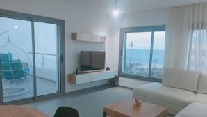 Een moderne woonkamer met een houten tafel, een bank en een televisie op een witte kast. Grote ramen bieden uitzicht op de zee en een balkon met een hangstoel.