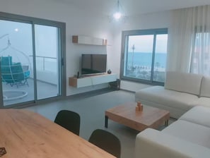 Ein modernes Wohnzimmer mit einem Holztisch, einer Couch und einem Fernseher auf einem weißen Schrank. Große Fenster bieten Blick auf das Meer und einen Balkon mit einer Hängematte.