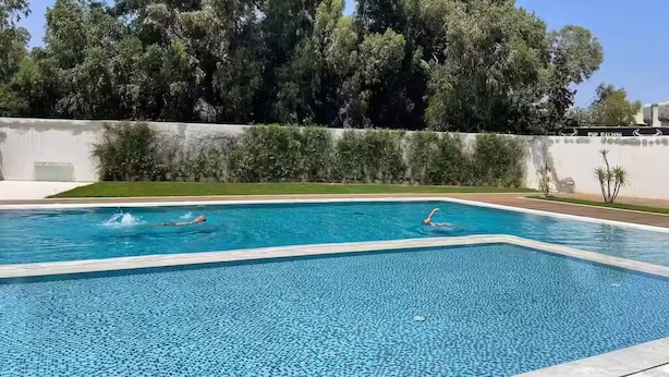 Een rechthoekig zwembad met helderblauw water, omringd door een lage witte muur en weelderige groene bomen op de achtergrond.