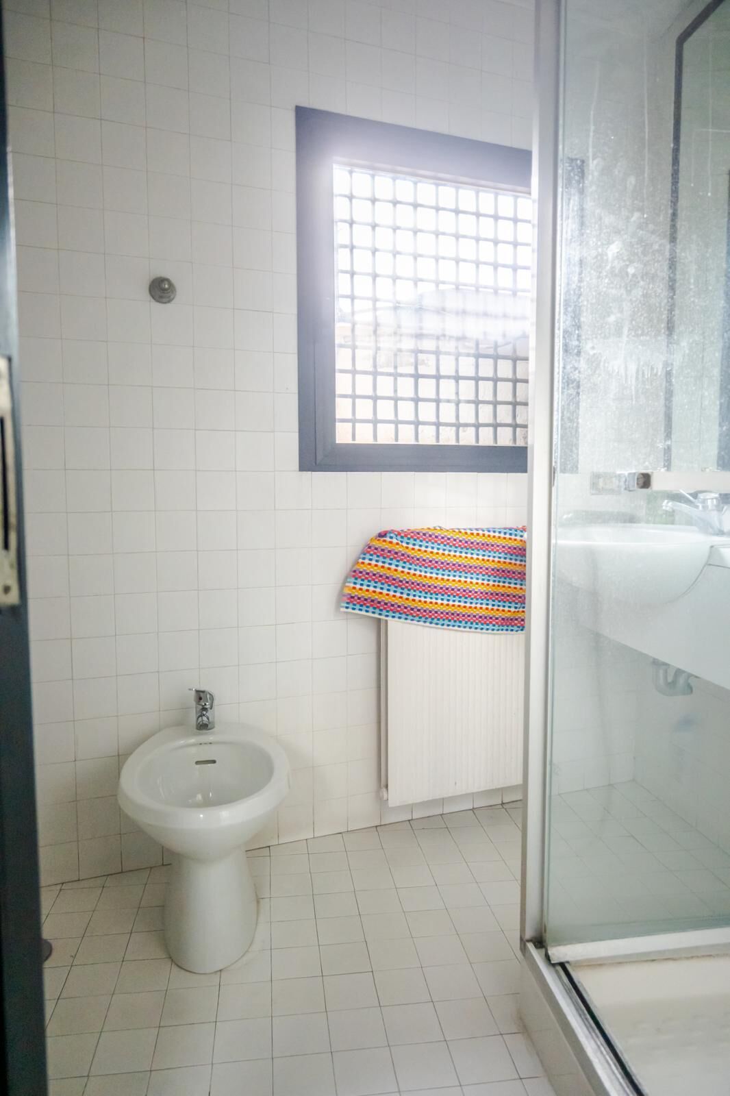 A bathroom with a bidet, a window with a grid pattern, and a towel hanging on a radiator.