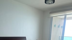 A bedroom with a large bed, a small table with a lamp, a chair, and a view of the ocean through a sliding door. - Oceánica Luxury Apartment (Salinas)