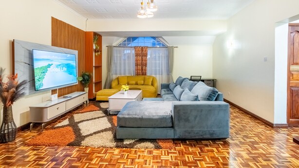 Ein modernes Wohnzimmer mit einem großen Flachbildfernseher, einem Sofa, einem gelben Sessel und einem gemusterten Teppich.