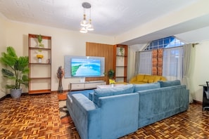 Ein Wohnzimmer mit blauem Sofa, Holzboden, Fernseher und einem Fenster mit Vorhängen.