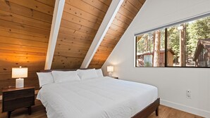 Une chambre avec un plafond en bois, un grand lit, deux tables de chevet avec des lampes et une fenêtre donnant sur des arbres et un chalet.