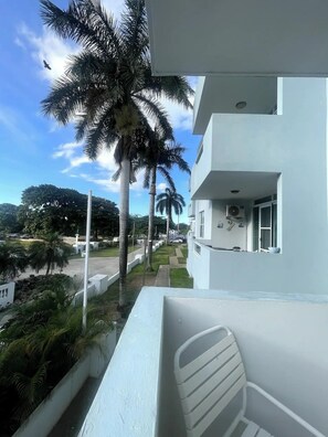 A balcony with a white chair, a white building with a fan and window, and palm trees in the background. - Spacious, Breezy 2 bdrm Condo! Great Location between Jobos Beach & Shacks Beach (Isabela)