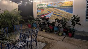 Una habitación con un mural de barcos en la playa, una mesa de vidrio con sillas y diversas plantas en macetas.