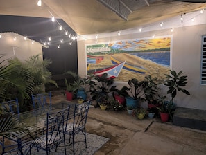 Una habitación con un mural de barcos en la playa, una mesa de vidrio con sillas y diversas plantas en macetas.