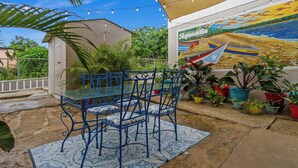 An outdoor patio with a blue metal table and chairs, surrounded by potted plants and a mural depicting a boat on a beach. - Unique 2-Home Family Getaway Near Beaches Airport Restaurants.  Ideal for Groups (Arenales)