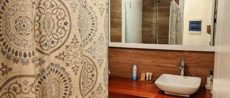 Un baño con cortina de ducha estampada, tocador de madera con lavamanos blanco y espejo sobre el lavamanos.