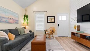 A living room with a grey sofa, a brown ottoman, a wicker chair, a TV on the wall, and a bookshelf. - Ocean Isle Retreat (Ocean Isle Beach)
