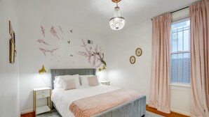 Un dormitorio con cama, mesitas de noche, una araña, una ventana con cortinas y adornos en la pared.
