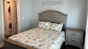 Een slaapkamer met een bed, voorzien van een bedlaken met een thema van de zee, een houten hoofdbord, een nachtkastje en een wandbordje met de tekst 'Relax, vakantietijd!'