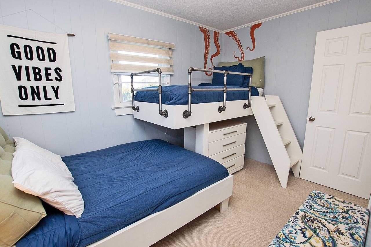 A bunk bed room with a bunk bed, a lower bed, and a ladder. There is a window with blinds, a door, and a wall with a sign that says "GOOD VIBES ONLY".