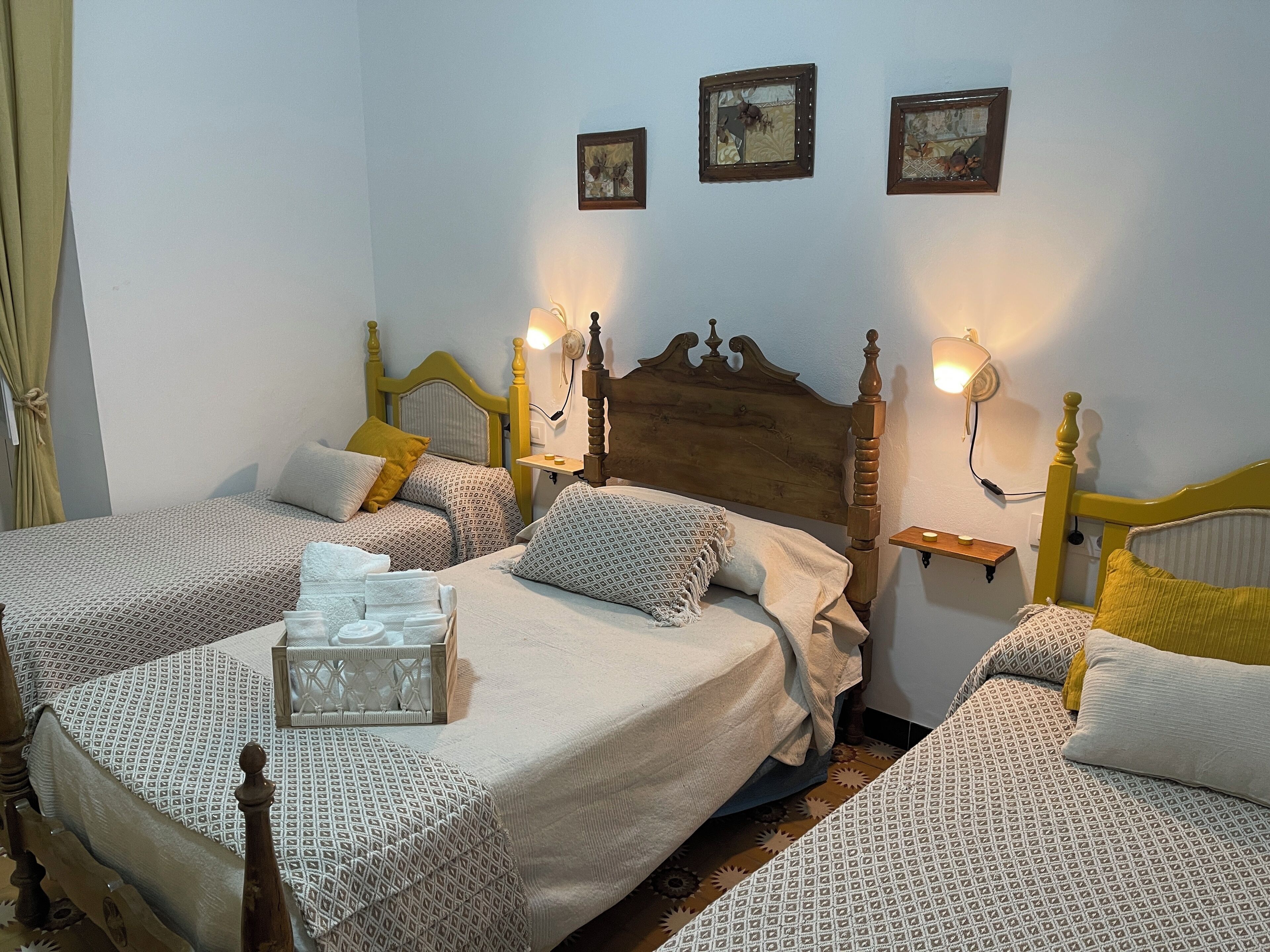 A room with two beds, each with patterned bedding and wooden headboards. There are wall-mounted lamps and a small shelf with a candle.