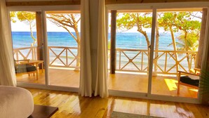 A room with a wooden floor, a large window with a view of the ocean, and a balcony with wooden furniture. - Turtle Beach Sanctuary Utila  (Isla de Utila)
