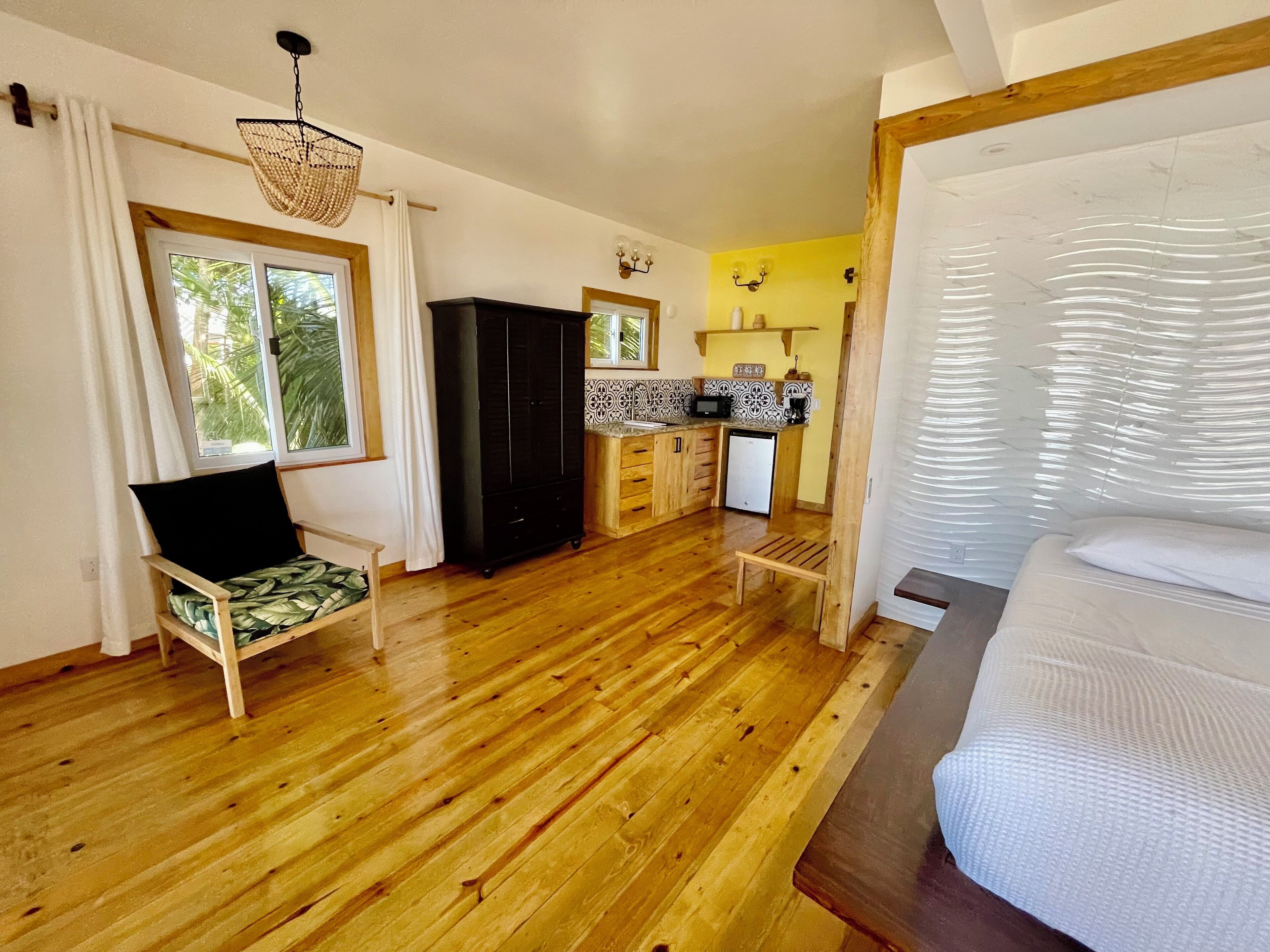 A cozy room with a wooden floor, a bed, a chair, a wardrobe, a small kitchen area, and a window with curtains.