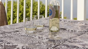 A table with a bottle and two glasses on a patterned tablecloth, set on a balcony with a white railing and a scenic view of a lake and mountains. - The Folly PLUS, University Place View Home with Pool, Perfect  for Celebrations. (University Place)