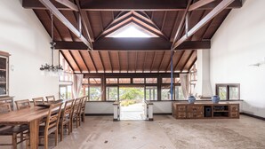 Une pièce spacieuse avec un plafond haut, des poutres apparentes, une grande table à manger et une vue par les fenêtres.