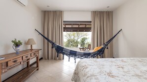 Ein Schlafzimmer mit Bett, Schreibtisch mit Schubladen, Lampe, Hängematte und Blick ins Freie.