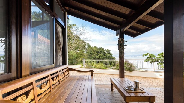 Eine hölzerne Bank und ein Tisch auf einer überdachten Terrasse mit Blick auf das Meer und viel Grün.