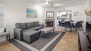 En moderne stue med en grå hjørnesofa, et sofabord og en pejs. Der er et spiseområde med et rundt bord og stole. Køkkenet ses i baggrunden med hvide skabe og hårde hvidevarer.
