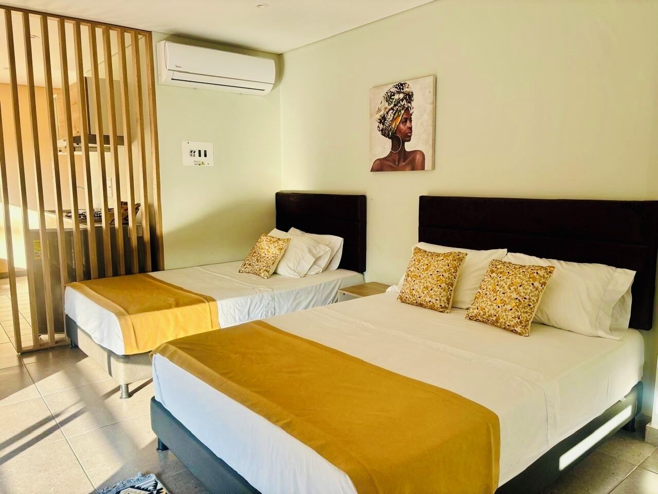 Habitación de hotel con dos camas, cada una con cabecero y almohadas. Se observa un aire acondicionado de pared y un cuadro sobre las camas.