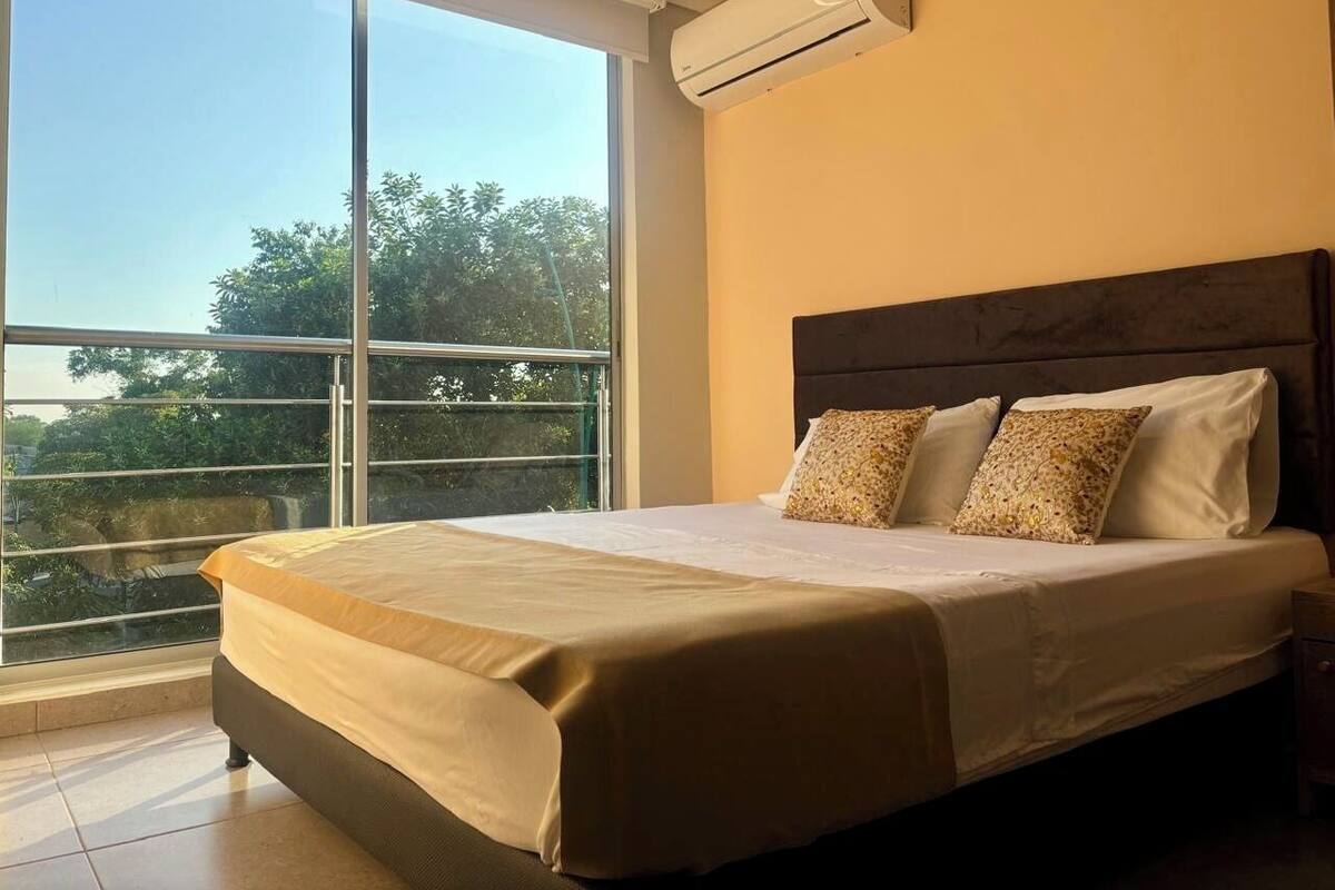 Habitación de hotel con un ventanal grande, cama con cabecera de madera y un equipo de aire acondicionado en la pared.