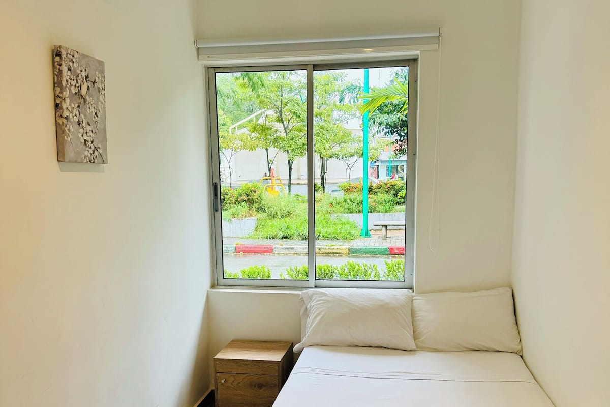 Una habitación con una cama, una mesita de noche de madera, un aire acondicionado instalado en la pared y una ventana que da a zonas verdes y una calle.