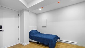 Ein kleines Zimmer mit einem Einzelbett, Holzboden, weißen Wänden und einer Deckenleuchte.