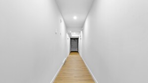 Un pasillo estrecho, bien iluminado, con paredes blancas y suelo de madera, que conduce a una puerta al final.