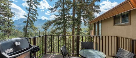 A deck with a grill, chairs, and a table overlooking a forest and mountains.
