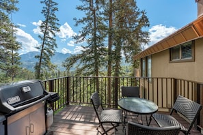 Eine Terrasse mit Grill, Stühlen und Tisch mit Blick auf einen Wald und Berge.