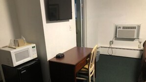 Ein kleines Hotelzimmer mit einem Holztisch, einer Mikrowelle, einem an der Wand befestigten Fernseher und einem Fenster mit Klimaanlage.