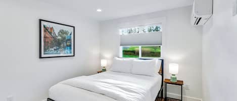Une chambre moderne avec un lit, deux tables de chevet avec des lampes, une fenêtre avec des stores, un climatiseur et un tableau encadré au mur.