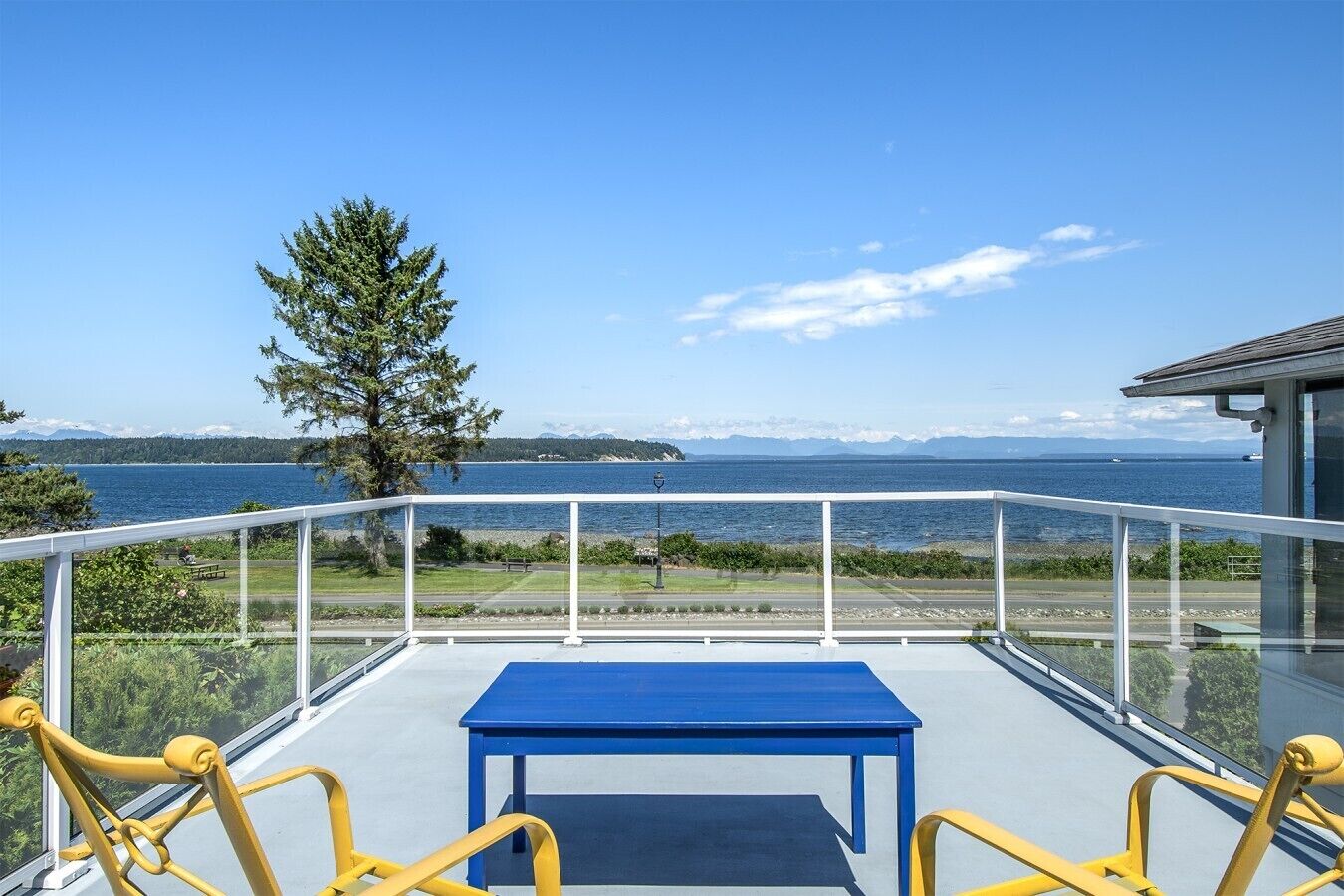 Balcón con una mesa azul y sillas amarillas, con vista a un cuerpo de agua y una costa distante.