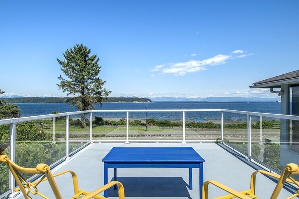A balcony with a blue table and yellow chairs, overlooking a body of water and a distant shoreline.