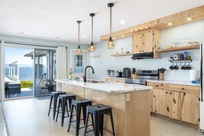A modern kitchen with a large island, bar stools, wooden cabinets, and a view of the sea.