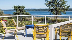 A balcony with yellow chairs and a table, overlooking a body of water and a forested landscape. - Oceanfront Central Seawalk Location | Patio, BBQ (Campbell River)