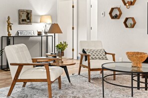 En stue med to hvide læderstole, et rundt glasbord, en lampe, et velkomstskilt og dekorative hexagonale hylder.