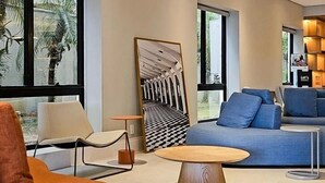 A modern living room with a large window, a blue sofa, an orange armchair, and a round wooden coffee table. - Suite Luxo a 5min do aerop de Congonhas (São Paulo)